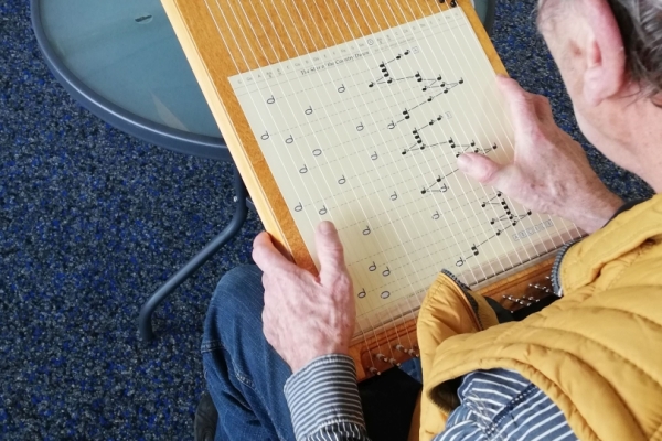 Bild: Ein Mann sitzt mit einer Harfe auf einem Stuhl. Unter den Saiten der Harfe liegt ein Blatt, das anzeigt, wo man an der Saiten zupfen muss.