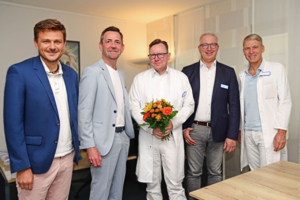 Fünf Männer stehen in einem Raum, einer hält einen Blumenstrauß