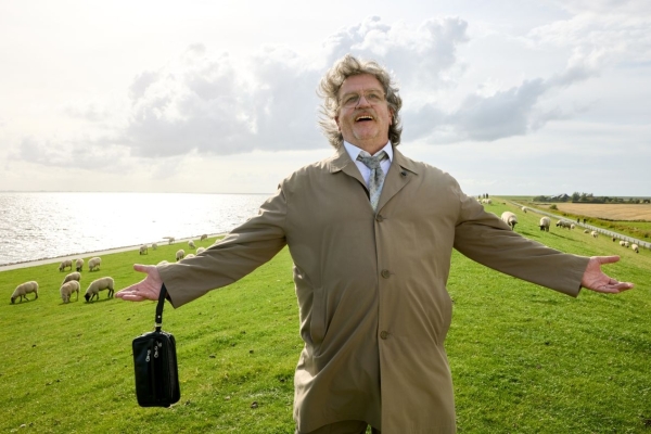 Bild: Horst Schlämmer steht mit ausgebreiteten Armen und mit einem Lächeln auf einem Deich. Im Hintergrund grasen Schafe. 