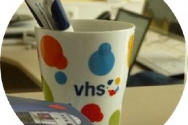 Bild: Eine Tasse mit der Aufschrift VHS und einigen Stiften