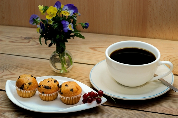 Bild: Eine Tasse Kaffee steht neben einem Teller mit drei Muffins. Dahinter steht eine kleine Vase mit einigen Blumen.