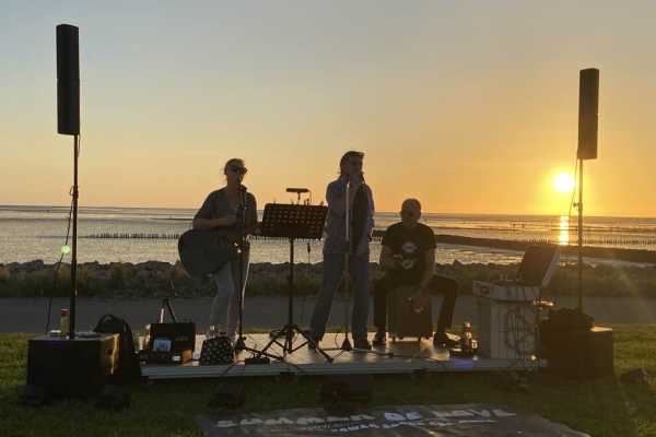 Bild: Die Band steht auf einer Bühne am Meer im Sonnenuntergang