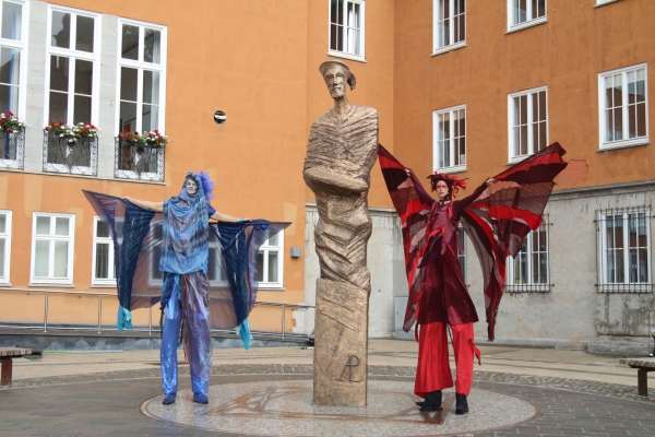 Bild: Zwei Stelzenläufer stehen rechts und links neben der Statue Allessandro Pasqualini auf dem Marktplatz