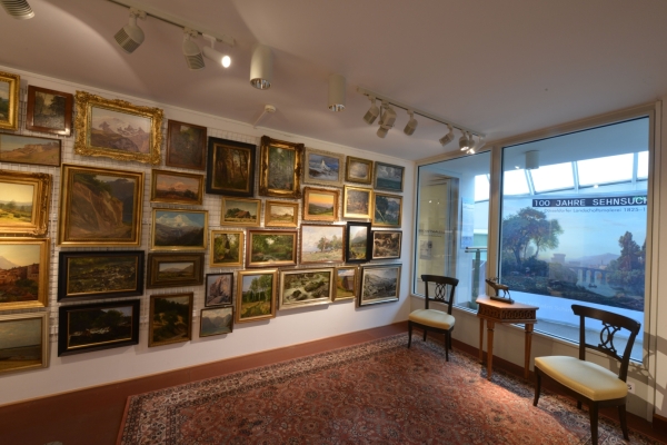 Bild: Die Ecke eines Zimmers. Ander einen Wand hängen viele Bilder nebeneinander. An der anderen Seite stehen 2 Stühle und ein Tisch.