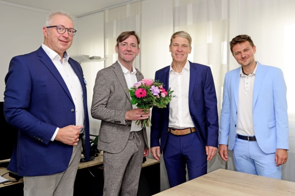 vier Männer stehen in einem Büro, einer mit Blumenstrauß