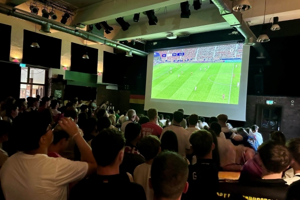 Bild: Auf einer Leinwand läuft ein Fußballspiel. Davor sitzen die Fans.
