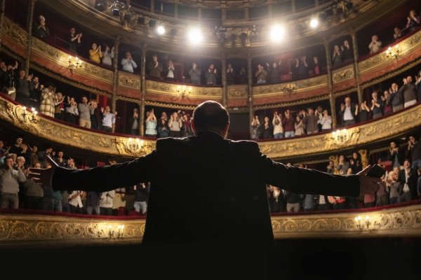 Bild: Ein Mann (wird von hinten gezeigt) steht mit weit ausgestreckten Armen auf der Bühne in einem Theater. Das Publikum macht 