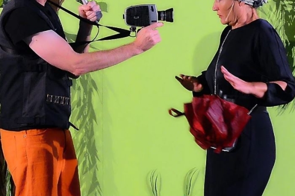 Spex! Ein Planet muss in die Werkstatt! Bild: Eine Mann (l) und eine Frau stehen sich gegenüber vor einem bunten Hintergrund. Der Mann interviewt ud filmt die Frau.