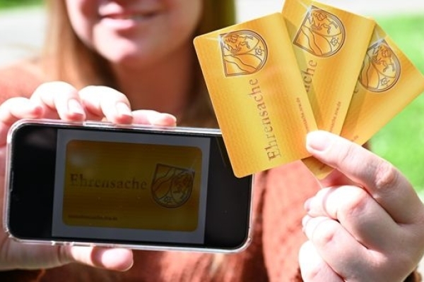 Bild: In GroÃŸaufanhme wird ein Smartphone mit einem Bild der App gezeigt sowie mehrere Ehrenamtskarten. Beides wird von einer Frau in die Kamera gehalten, die im HIntergrund angedeutet zu sehen ist..