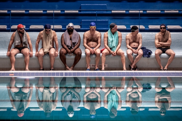 Bild: 7 MÃ¤nner in Badekleidung sitzen am Ran des Schwimmbeckens auf einer Bank.