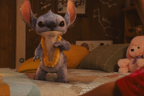 Kino: Lilo & Stitch Bild: Stitch steht auf einem Bett mit Kissen und Stofftier. Einige Lampen erhellen ein wenig das Zimmer.