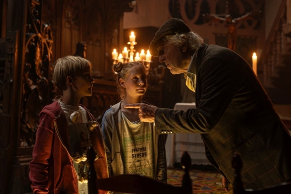Kino: Max und die wilde 7: Die Geister-Oma Bild: Ein Junge und ein Mädchen stehen in einem dunklen, von Kerzen erhellten Raum. Vor ihnen steht vornübergebeugt ein alter Mann, der mit dem Finger auf sie zeigt.