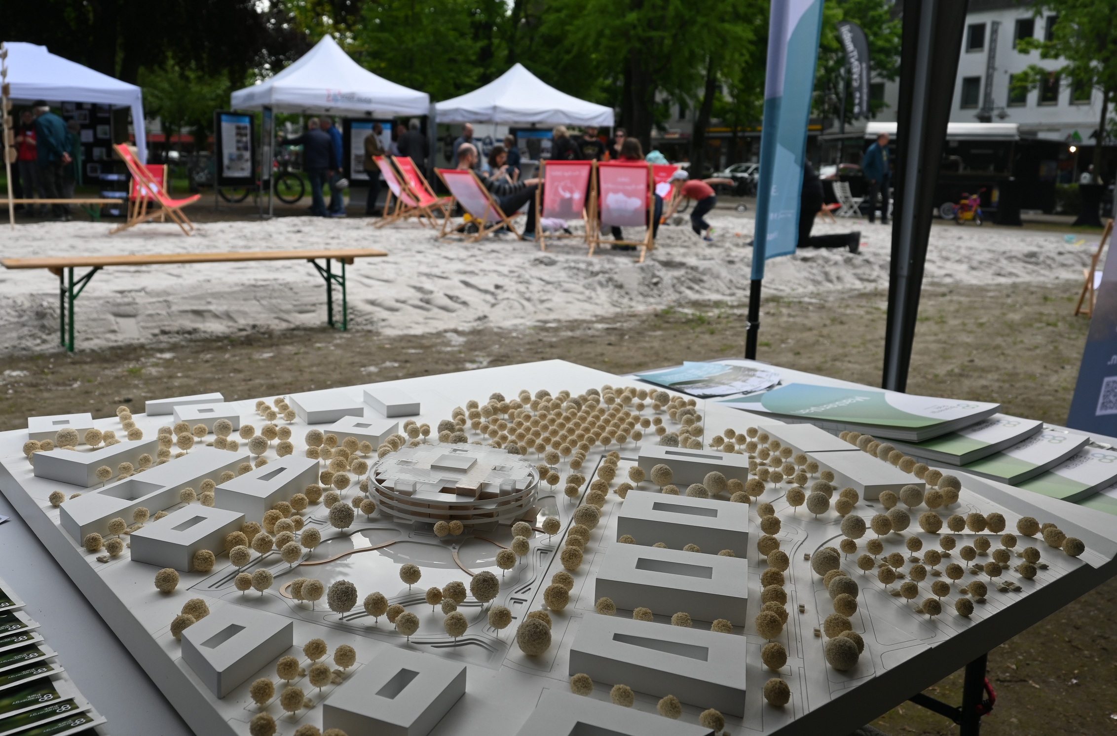 Im Vordergrund ein Modell - Wie könnte Jülich in Zukunft aussehen - im Hintergrund sitzen einige Menschen in Liegestühlen und Kinder spielen im Sand. Dahinter kleine wei�?e Pavillons mit Infoständen.